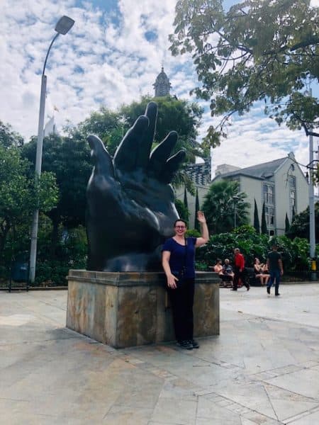 woman waving in front of an open hand statue, places to go in colombia, colombia, colombia destinations, medellin, things to do in medellin, what to do in medellin, botero, botero square, botero plaza, rafael uribe palace of culture