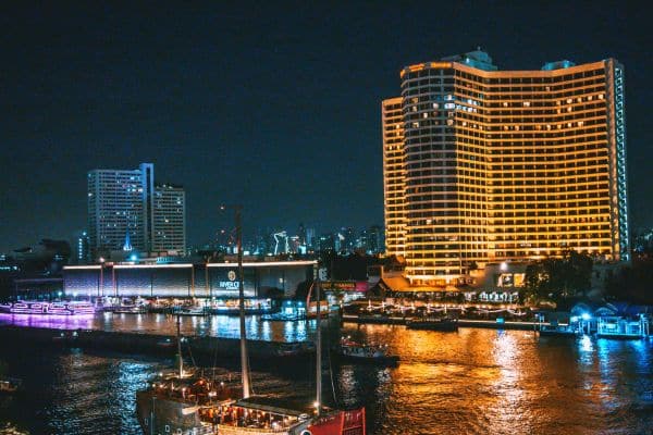 riverfront district at night, tall hotel in the distance, boats in the water, best hotels to stay in bangkok, places to stay in bangkok, best places to stay in bangkok