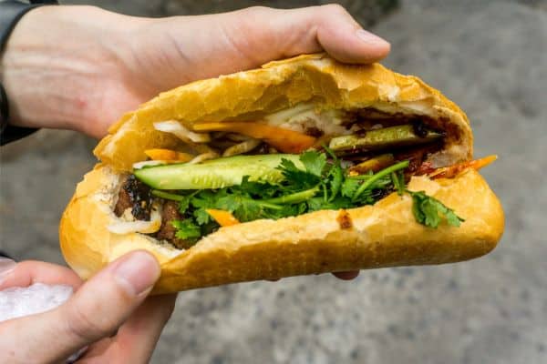 men holding a banh mi sandwich up close so you can see everything inside, banh mi filled with cucumbers, carrots, meat, and cilantro