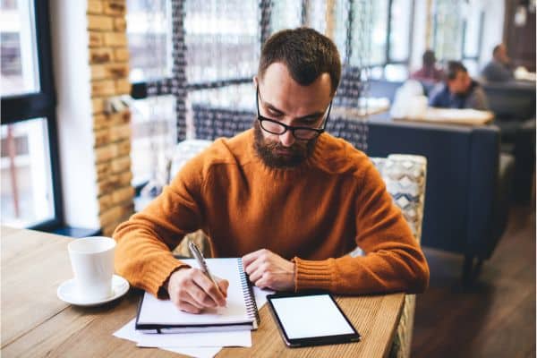 man sitting in a coffee shop writing in a notebook, side jobs from home, remote side hustles, side hustle jobs, online hustles