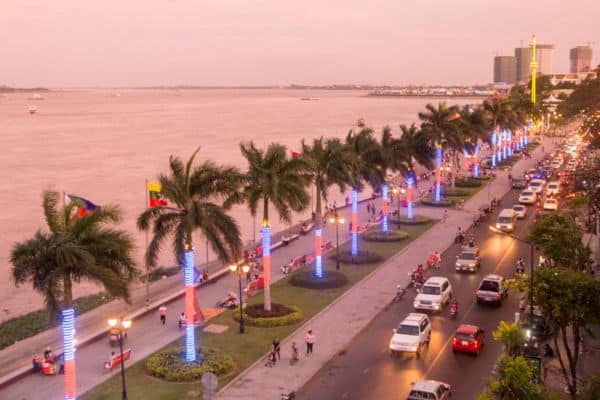 sisowath quay at sunset, lights around the palm trees, cats going up and down the roads, river and tall buildings in the distance