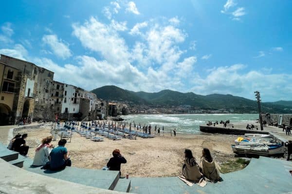 people sitting on steps near the sand, ocean and mountains in teh distance, city buildings on the left side, blue and clear sky with a few clouds