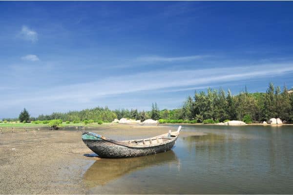beach at vung tau, small boat sitting in the water, trees along the shoreline with clear and blue skies in the back, day trip fro ho chi minh