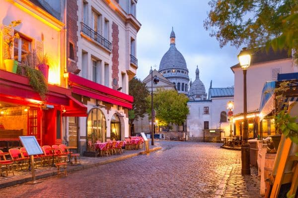 montmarte neighbrohood paris, top of basilica in the background, cobblstone old street with restaurants and outdoor dining on both sides